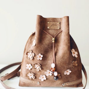 Peut inclure: Un sac seau en daim marron avec une fermeture &agrave; cordon et une longue bandouli&egrave;re. Le sac est d&eacute;cor&eacute; de fleurs en cuir rose et porte une petite &eacute;tiquette en cuir marron avec le mot "folks" &eacute;crit dessus.