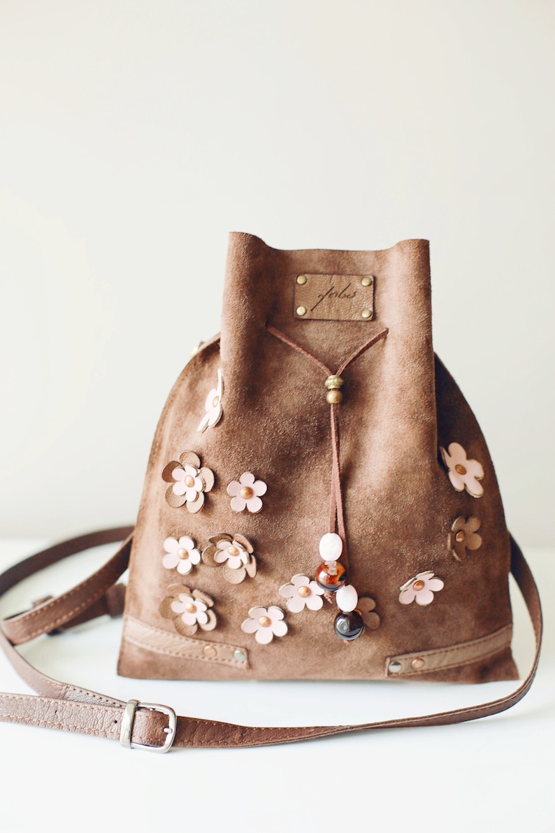 Peut inclure: Un sac seau en daim marron avec une fermeture &agrave; cordon et une longue bandouli&egrave;re. Le sac est d&eacute;cor&eacute; de fleurs en cuir rose et porte une petite &eacute;tiquette en cuir marron avec le mot "folks" &eacute;crit dessus.