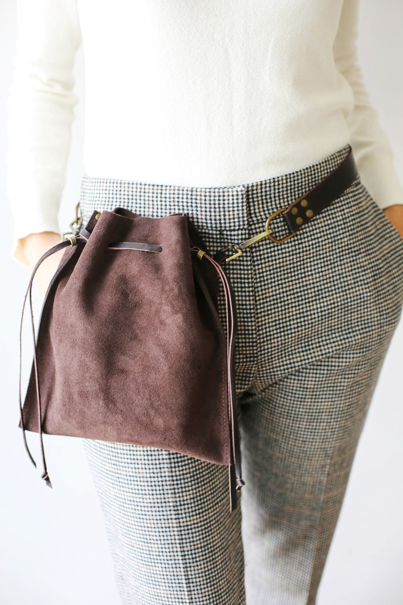May include: A brown suede drawstring bag with a leather strap attached to a brown leather belt. The bag is worn on the side of the body and has a long strap that hangs down.