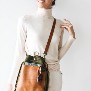 May include: A medium-sized crossbody bag with a brown leather front pocket and a green suede body. The bag has a brown leather strap and a zipper closure. Sunglasses are tucked into the front pocket. The text "Medium SIZE Crossbody Style" is at the top.