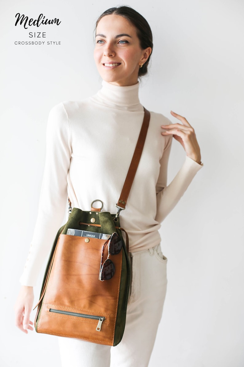 May include: A medium-sized crossbody bag with a brown leather front pocket and a green suede body. The bag has a brown leather strap and a zipper closure. Sunglasses are tucked into the front pocket. The text "Medium SIZE Crossbody Style" is at the top.