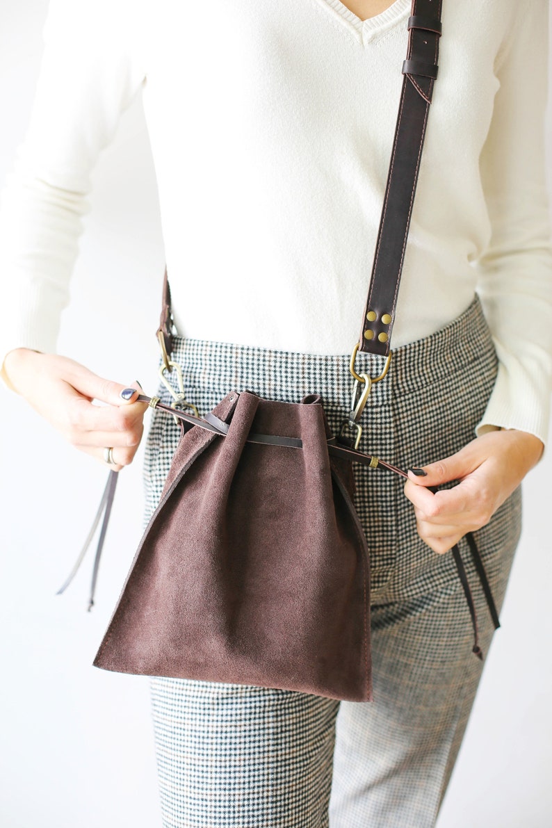 May include: A brown suede drawstring bag with a long brown leather strap. The bag is worn over the shoulder of a person wearing a white sweater and plaid pants.