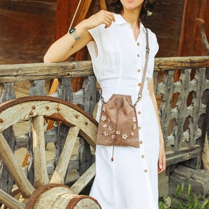 Peut inclure: Une femme portant une robe blanche avec un sac en daim brun orn&eacute; de fleurs. Elle se tient devant une roue en bois et un b&acirc;timent en bois.