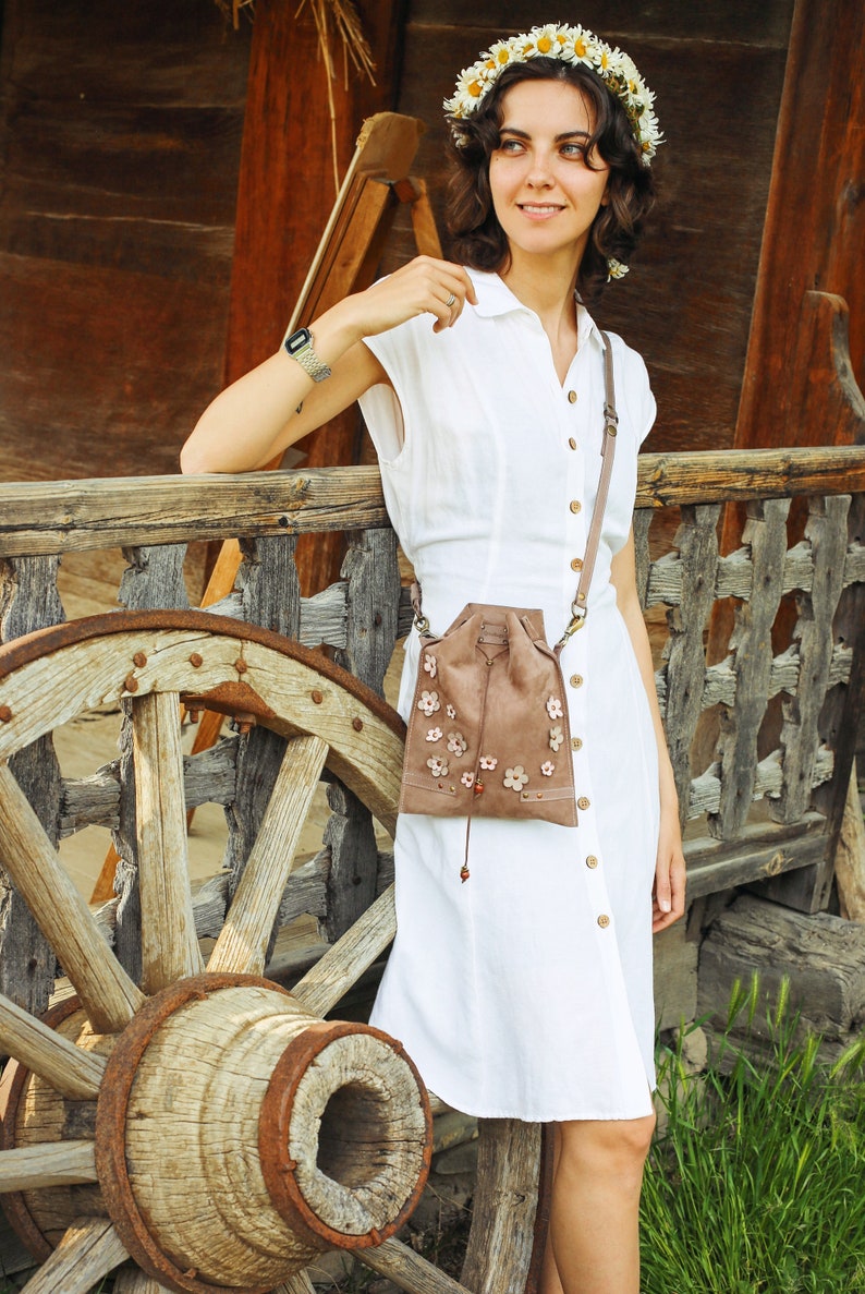 Peut inclure: Une femme portant une robe blanche avec un sac en daim brun orn&eacute; de fleurs. Elle se tient devant une roue en bois et un b&acirc;timent en bois.