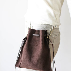 May include: A brown suede drawstring bag with a long strap attached to a belt loop. The bag is worn on the hip of a person wearing white pants and a white sweater.