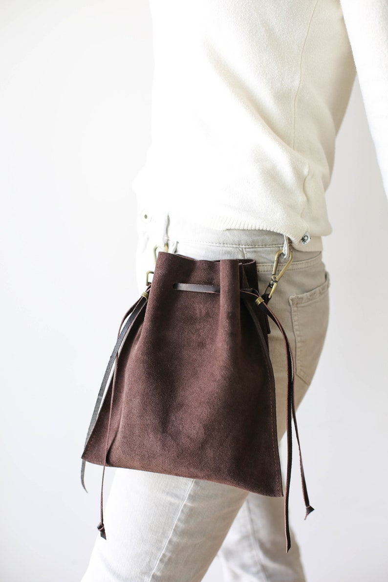 May include: A brown suede drawstring bag with a long strap attached to a belt loop. The bag is worn on the hip of a person wearing white pants and a white sweater.