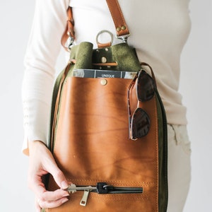 May include: A brown and green leather backpack with a drawstring closure and a front zipper pocket. The bag has a brown leather strap and a pair of sunglasses hanging from it. The word "UNIQUE" is visible on a magazine inside the bag.