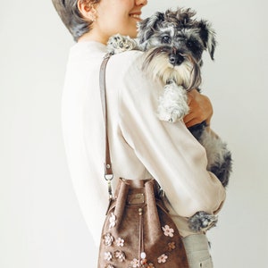 Peut inclure: Une femme aux cheveux courts bruns tient un petit chien gris et blanc dans ses bras. Elle porte une chemise blanche et un jean brun clair. Elle porte &eacute;galement un sac seau en daim brun avec des embellissements de fleurs roses.