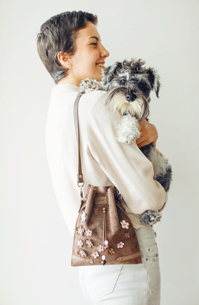 Peut inclure: Une femme aux cheveux courts bruns tient un petit chien gris et blanc dans ses bras. Elle porte une chemise blanche et un jean brun clair. Elle porte &eacute;galement un sac seau en daim brun avec des embellissements de fleurs roses.