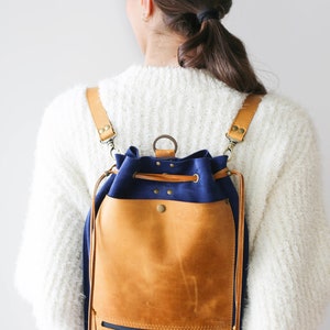 May include: A blue and brown leather backpack with a drawstring closure and a zippered pocket on the front. The backpack has adjustable straps and is worn by a person.