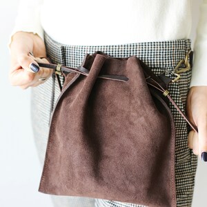 May include: A brown suede drawstring bag with a leather strap and brass hardware. The bag is worn over the waist.