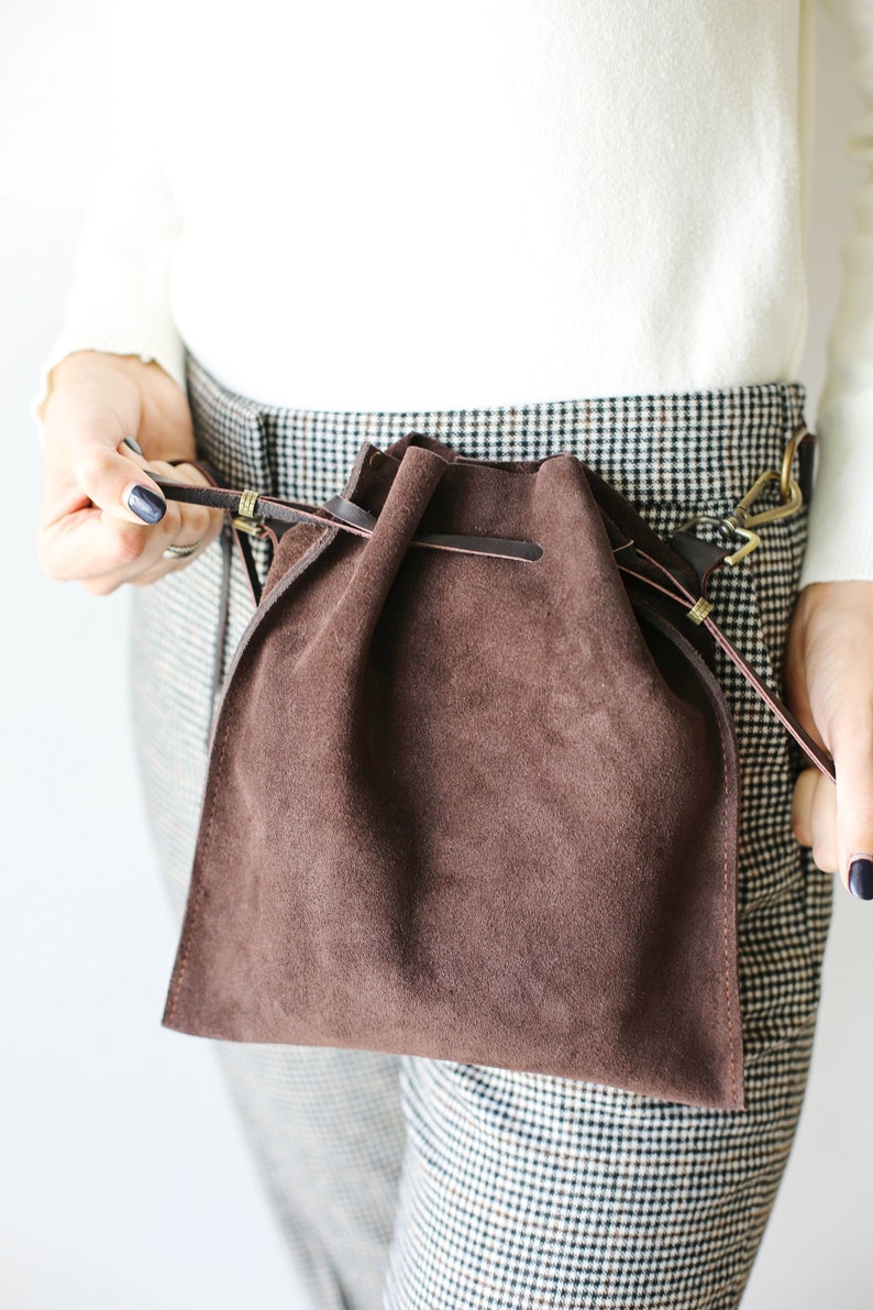 May include: A brown suede drawstring bag with a leather strap and brass hardware. The bag is worn over the waist.