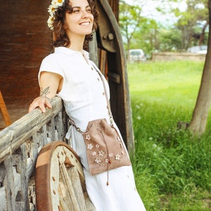 Peut inclure: Une femme portant une robe blanche et une couronne de fleurs de marguerite se tient &agrave; c&ocirc;t&eacute; d'une roue en bois. Elle porte un sac en daim brun avec des broderies florales.