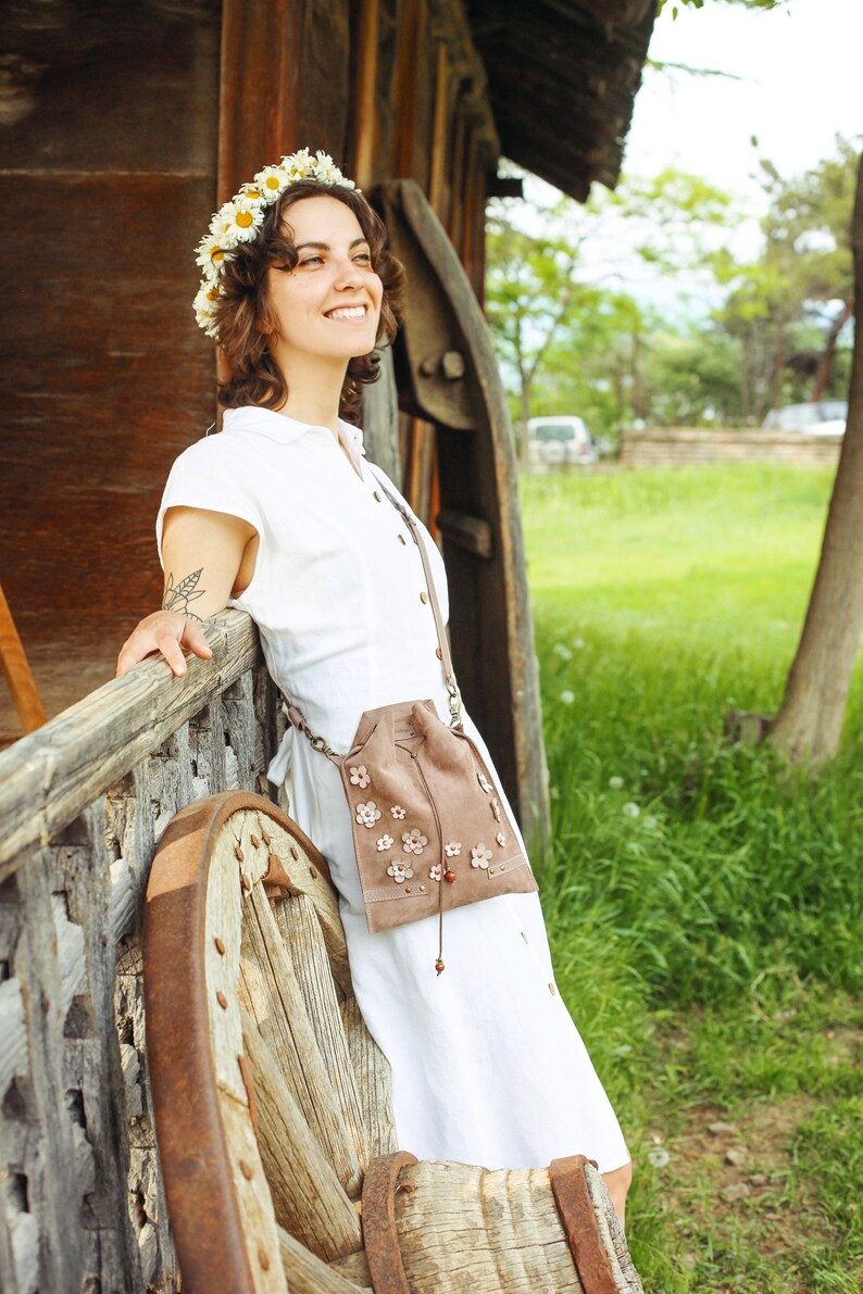 Peut inclure: Une femme portant une robe blanche et une couronne de fleurs de marguerite se tient &agrave; c&ocirc;t&eacute; d'une roue en bois. Elle porte un sac en daim brun avec des broderies florales.