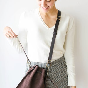 May include: A brown suede drawstring bag with a long brown leather strap. The bag is worn over the shoulder of a person wearing a white sweater and black and white checkered pants.