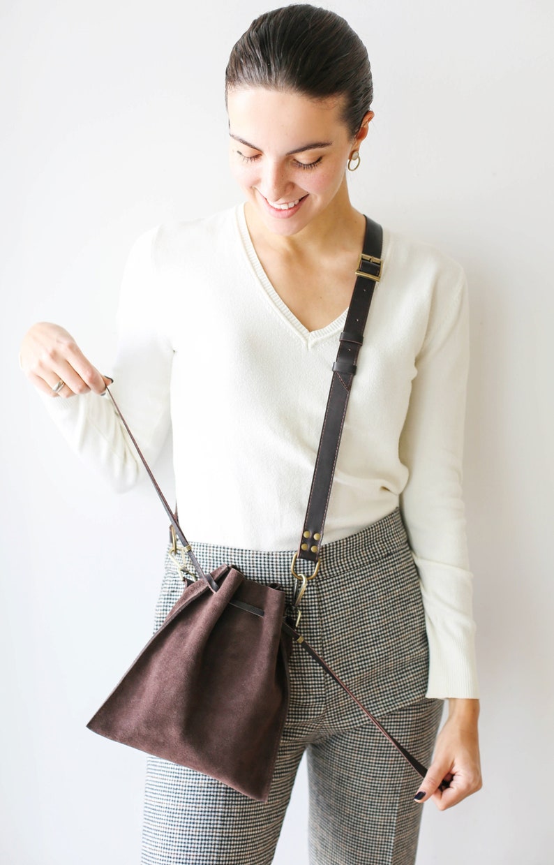 May include: A brown suede drawstring bag with a long brown leather strap. The bag is worn over the shoulder of a person wearing a white sweater and black and white checkered pants.