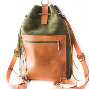 May include: A green suede and brown leather backpack with adjustable straps. The backpack features a drawstring closure, a front zippered pocket, and a top handle. The straps and accents are a lighter shade of brown.
