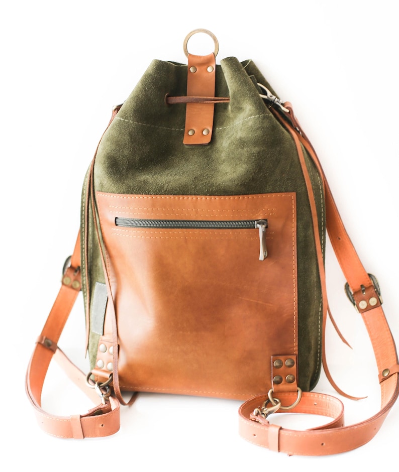 May include: A green suede and brown leather backpack with adjustable straps. The backpack features a drawstring closure, a front zippered pocket, and a top handle. The straps and accents are a lighter shade of brown.