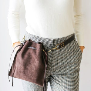May include: A brown suede drawstring bag with a leather strap attached to a brown leather belt. The bag is worn on the side of a person wearing a white sweater and black and white checkered pants.