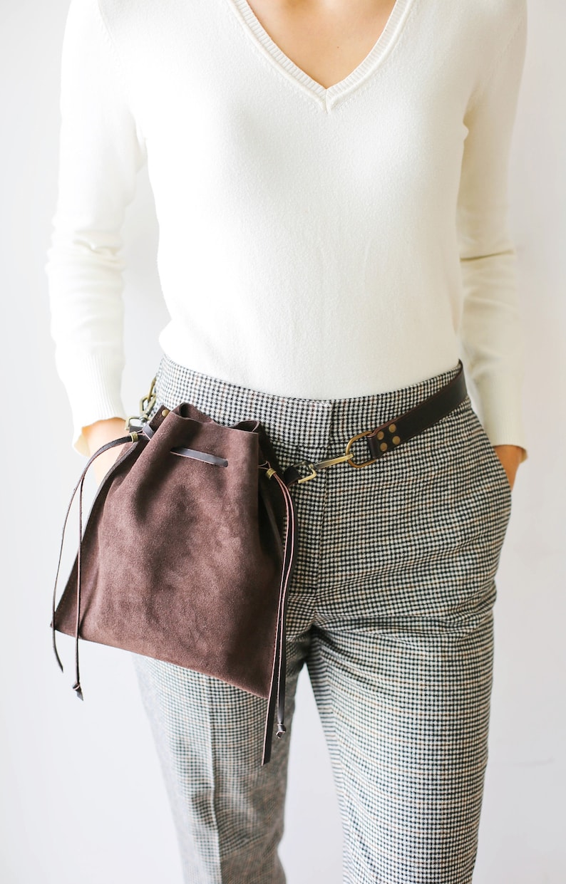 May include: A brown suede drawstring bag with a leather strap attached to a brown leather belt. The bag is worn on the side of a person wearing a white sweater and black and white checkered pants.