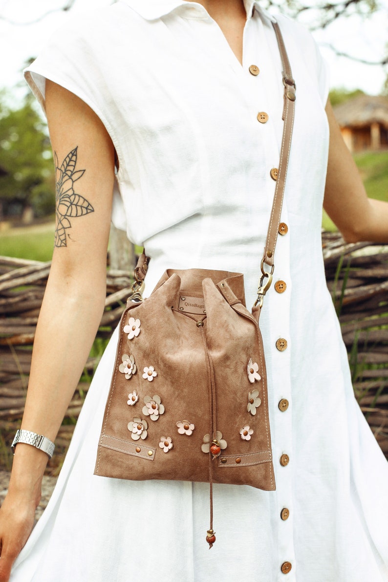 Peut inclure: Un sac seau en daim marron avec une fermeture &agrave; cordon et une longue bandouli&egrave;re. Le sac est d&eacute;cor&eacute; d'applications de fleurs roses et porte une petite &eacute;tiquette en cuir marron avec le texte "Ossa House".
