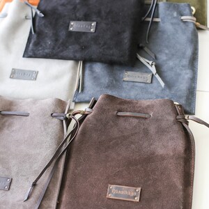 May include: A collection of six suede drawstring bags in various colors, including brown, gray, black, and green. Each bag features a leather label with the text "OilsBags".