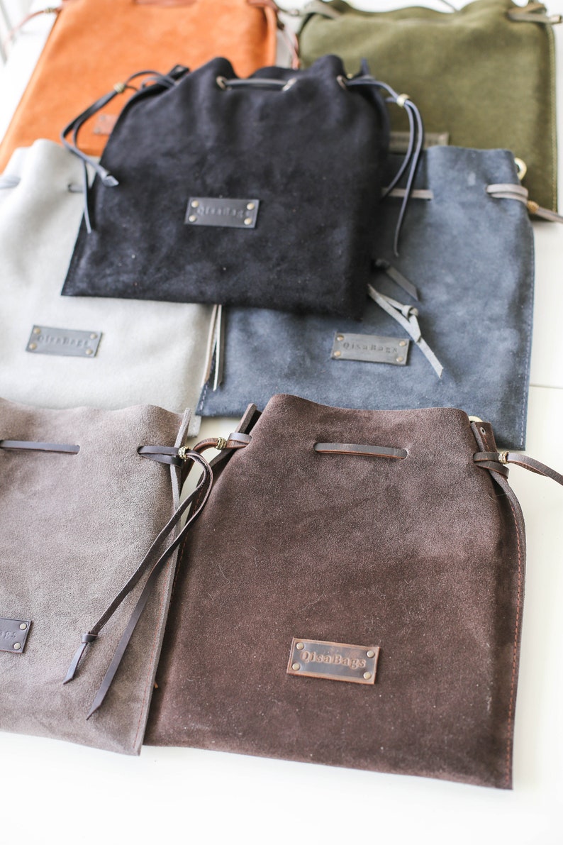 May include: A collection of six suede drawstring bags in various colors, including brown, gray, black, and green. Each bag features a leather label with the text "OilsBags".