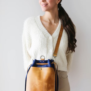 May include: A tan leather bucket bag with a blue canvas bottom. The bag has a drawstring closure and a zipper pocket on the front. The bag is worn on a shoulder strap.