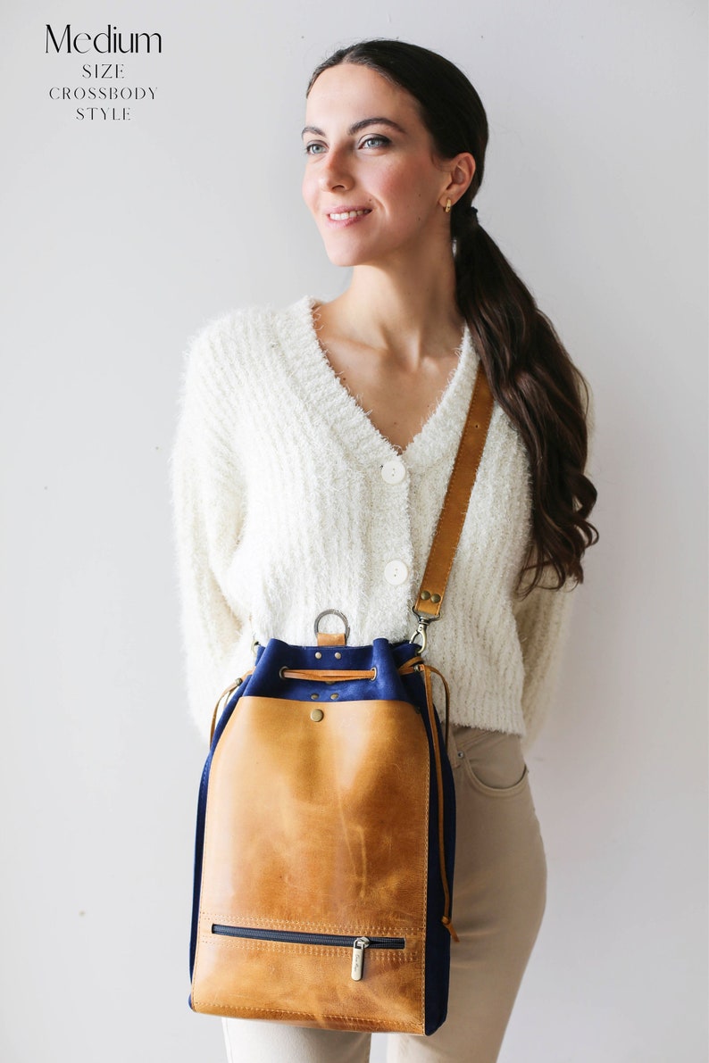 May include: A tan leather bucket bag with a blue canvas bottom. The bag has a drawstring closure and a zipper pocket on the front. The bag is worn on a shoulder strap.