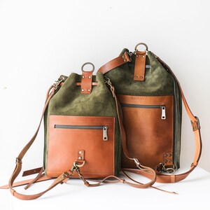 May include: Two olive green and brown leather backpacks. The backpacks have a drawstring closure, a front pocket with a zipper, and adjustable straps. The bags have a metal ring at the top and are made of suede and leather.