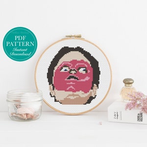 May include: A cross-stitch pattern of a person's face with a red mask. The pattern is on a round wooden frame. The frame is sitting on a white surface.