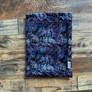 Puede incluir: Un artículo de tela rectangular con fondo negro, que presenta un estampado de hojas azules y moradas y estampado de leopardo blanco. El artículo está doblado y tiene una pequeña etiqueta blanca con texto.