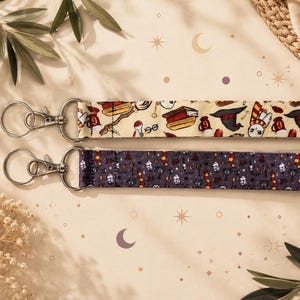 Peut inclure: Deux cordons porte-clés à motifs avec fermoirs en métal argenté. L'un est de couleur crème avec un motif répété de livres, de chapeaux et d'autres icônes. L'autre est violet foncé avec un motif similaire.
