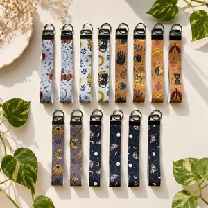 Op de afbeelding: Een collectie van veertien stoffen polsband-sleutelhangers met diverse prints, waaronder motten, maansikkels, hemelse motieven, botanische bladeren en katten, elk bevestigd aan een zwarte metalen sluiting en een ronde metalen ring.
