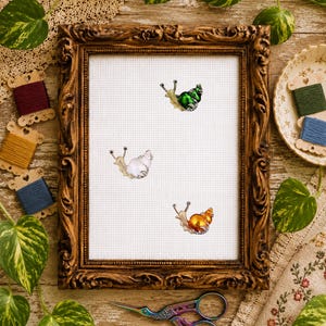May include: A white cross-stitch fabric piece framed in an ornate dark wood frame, featuring three embroidered snails with glossy shells in metallic green, pearlescent white, and vibrant orange, positioned with iridescent metal scissors below.