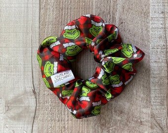 Puede incluir: Un scrunchie de cuadros rojos y negros con un estampado de cara verde del Grinch. El scrunchie tiene una etiqueta blanca que dice "OUTLAW STITCHING".
