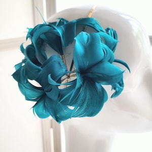 Puede incluir: Tocado de plumas turquesas, exhibido sobre una cabeza de maniquí blanca. El tocado presenta una disposición circular de plumas turquesas grandes y vibrantes. Las plumas están agrupadas, creando un diseño voluminoso y llamativo. La diadema es dorada.