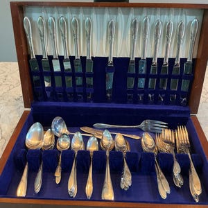 May include: A wooden storage chest with a blue velvet interior containing a silver-toned flatware set. The set includes various knives, forks, and spoons arranged in slots and compartments, featuring simple, smooth handles and polished metal surfaces.