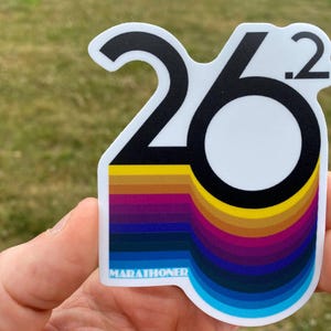 May include: A white sticker with the text "26.2" in black, with a rainbow gradient design below. The word "MARATHONER" is printed in white at the bottom. The sticker is held in a hand against a blurred green background.