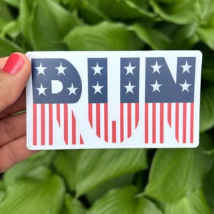 May include: A rectangular sticker with the word "RUN" in red, white, and blue. The letters are designed with a flag theme, featuring blue sections with white stars and red and white stripes.
