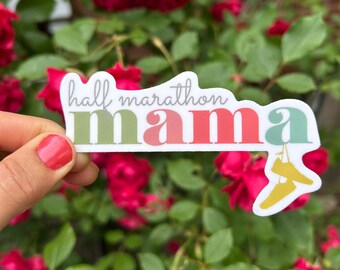 May include: A white sticker with the text "hall marathon mama" in a mix of colors. The word "mama" is in red, green, and pink. Two yellow sneakers are hanging from the word "mama". The sticker is held in front of a background of red roses and green leaves.