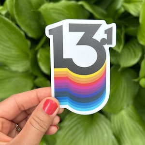 May include: A sticker featuring the number "13" in black and white with a rainbow-colored wave design at the bottom. The sticker is held against a backdrop of green leaves.