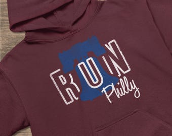 Peut inclure: Sweat à capuche bordeaux avec un motif blanc et bleu. Le motif présente le mot "RUN" avec une silhouette bleue de la Cloche de la Liberté et le mot "Philly" en écriture cursive.