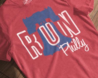 Peut inclure: T-shirt rouge avec un motif graphique représentant la Cloche de la Liberté en bleu et les mots "RUN Philly" en blanc. Le t-shirt a un col rond et des manches courtes. Le t-shirt est fait d'une matière douce.