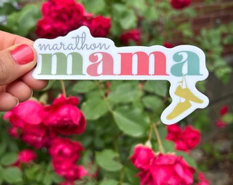 May include: A sticker with the words "marathon mama" in a colorful, retro font. The word "mama" is in green, pink, and teal. A pair of yellow running shoes is on the right side of the sticker. The background is a rose bush with red flowers.