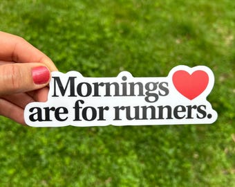 May include: A white sticker with the text "Mornings are for runners." in black, with a red heart symbol. The sticker is held against a background of green grass.