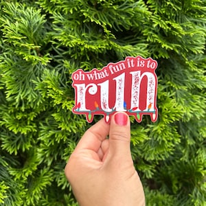May include: A red sticker with the words "oh what fun it is to RUN" in white, decorated with colorful string lights. The sticker is held in a hand against a backdrop of green foliage.