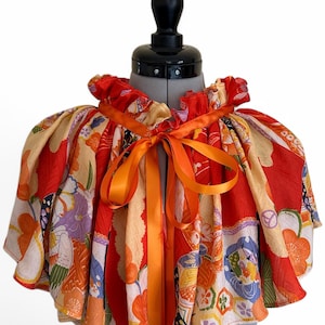 Könnte beinhalten: Ein kurzes Cape mit einem lebhaften Blumenmuster in Rot, Orange und Creme mit violetten und grünen Akzenten. Das Kleidungsstück hat einen gerüschten Kragen und wird vorne mit einem leuchtend orangefarbenen Satinband zu einer Schleife gebunden.