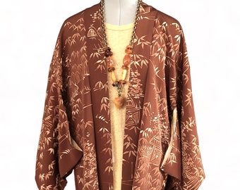 May include: A brown kimono-style jacket with a bamboo and architectural pattern, worn over a yellow top. A long necklace with amber and brown beads hangs from the neckline. The jacket is displayed on a black mannequin.
