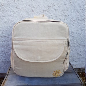 May include: Beige backpack with a flap pocket and a top handle. The bag has a zipper closure and a small embroidered sun design in yellow on the lower right corner. The backpack is made of a textured fabric.